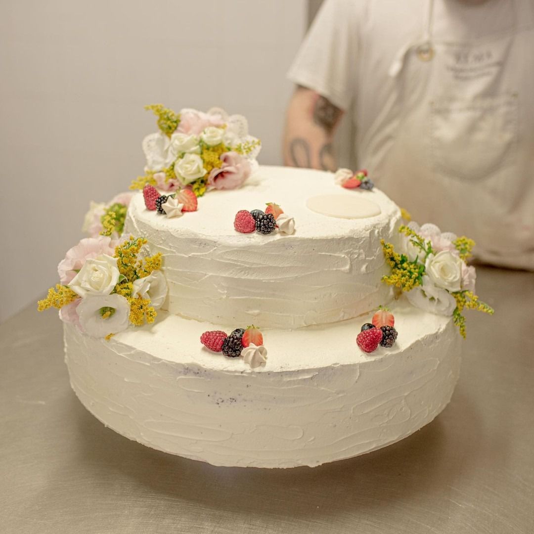 Torta bianca a due piani decorata con fiori e bacche, su una superficie metallica, con il fornaio sullo sfondo.