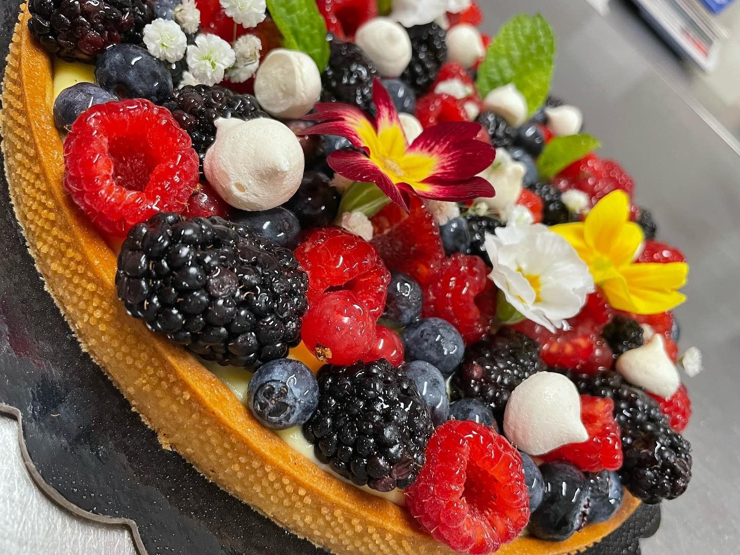 Crostata ai frutti di bosco, ricca di lamponi, more, mirtilli, meringhe e fiori commestibili, su una base scanalata.