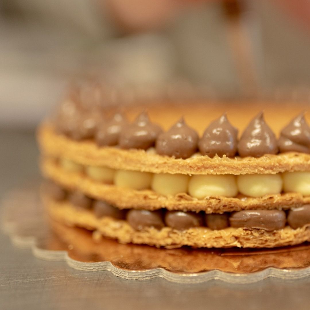 Primo piano di una pasta sfoglia a strati con ripieno di crema e glassa al cioccolato in superficie. La pasta sfoglia è servita su un piatto dorato con decorazioni ondulate.