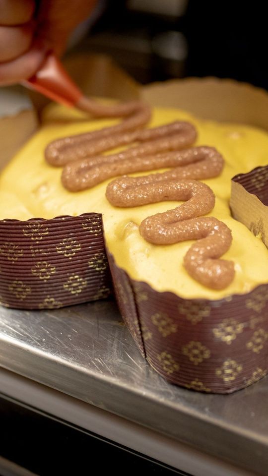 Una persona che stende la glassa marrone su una torta gialla avvolta in un pirottino di carta marrone, probabilmente in un ambiente da pasticceria.