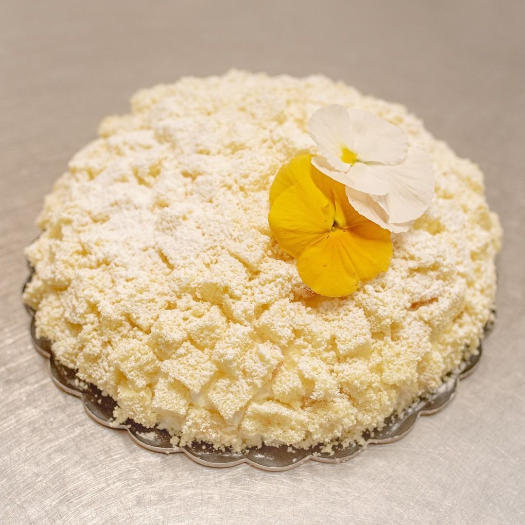 Una torta rotonda, ricoperta di briciole bianche e spolverata di zucchero a velo, è guarnita con fiori commestibili gialli e bianchi.