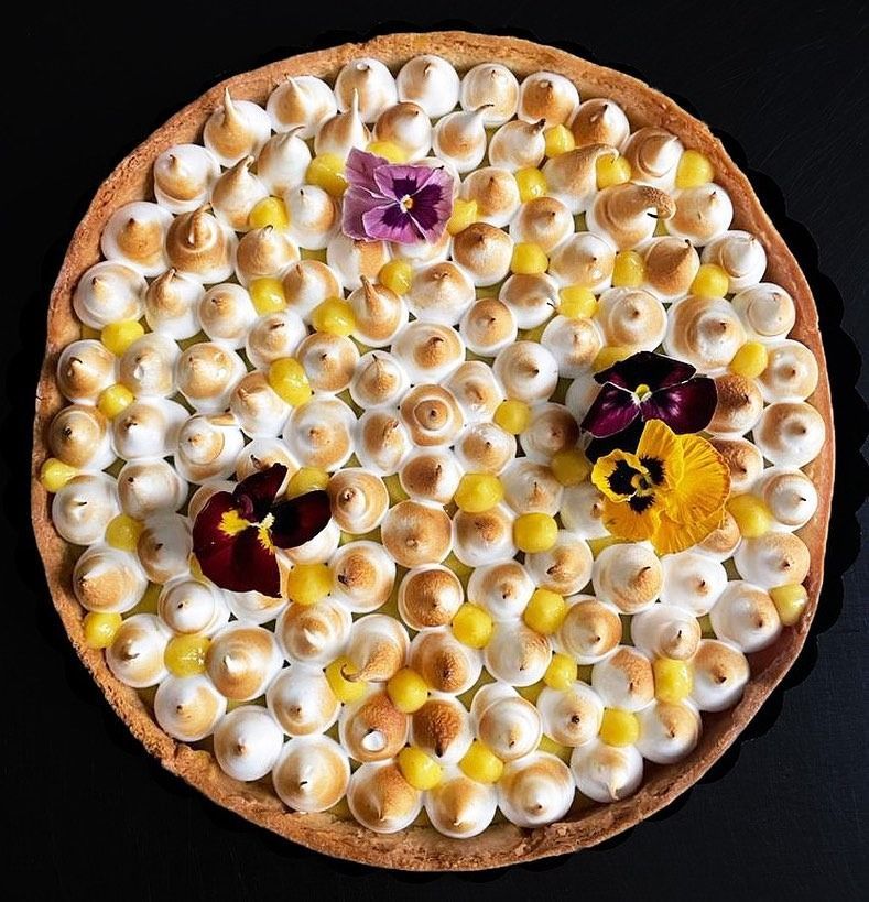 Crostata al limone e meringa ricoperta di meringa dorata, fiori commestibili e crema al limone su una superficie nera.