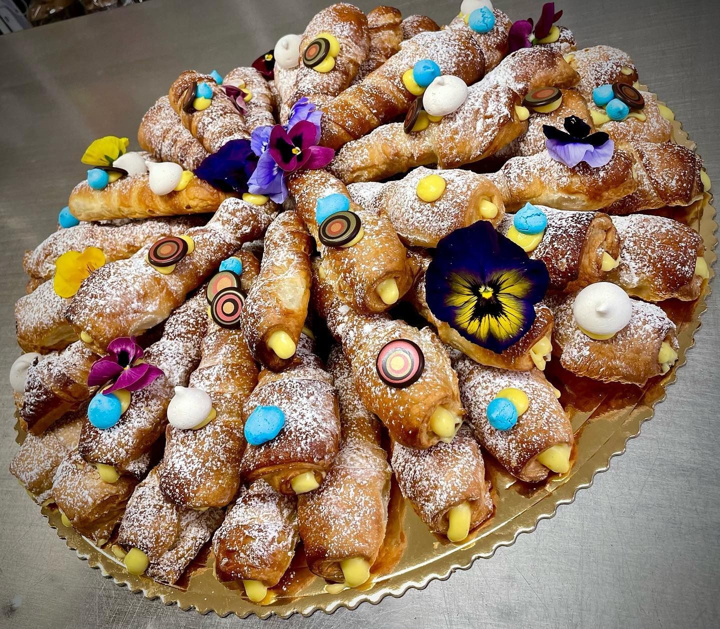 Un vassoio decorato con cannoli, spolverati con zucchero a velo e decorati con fiori commestibili e dettagli colorati.
