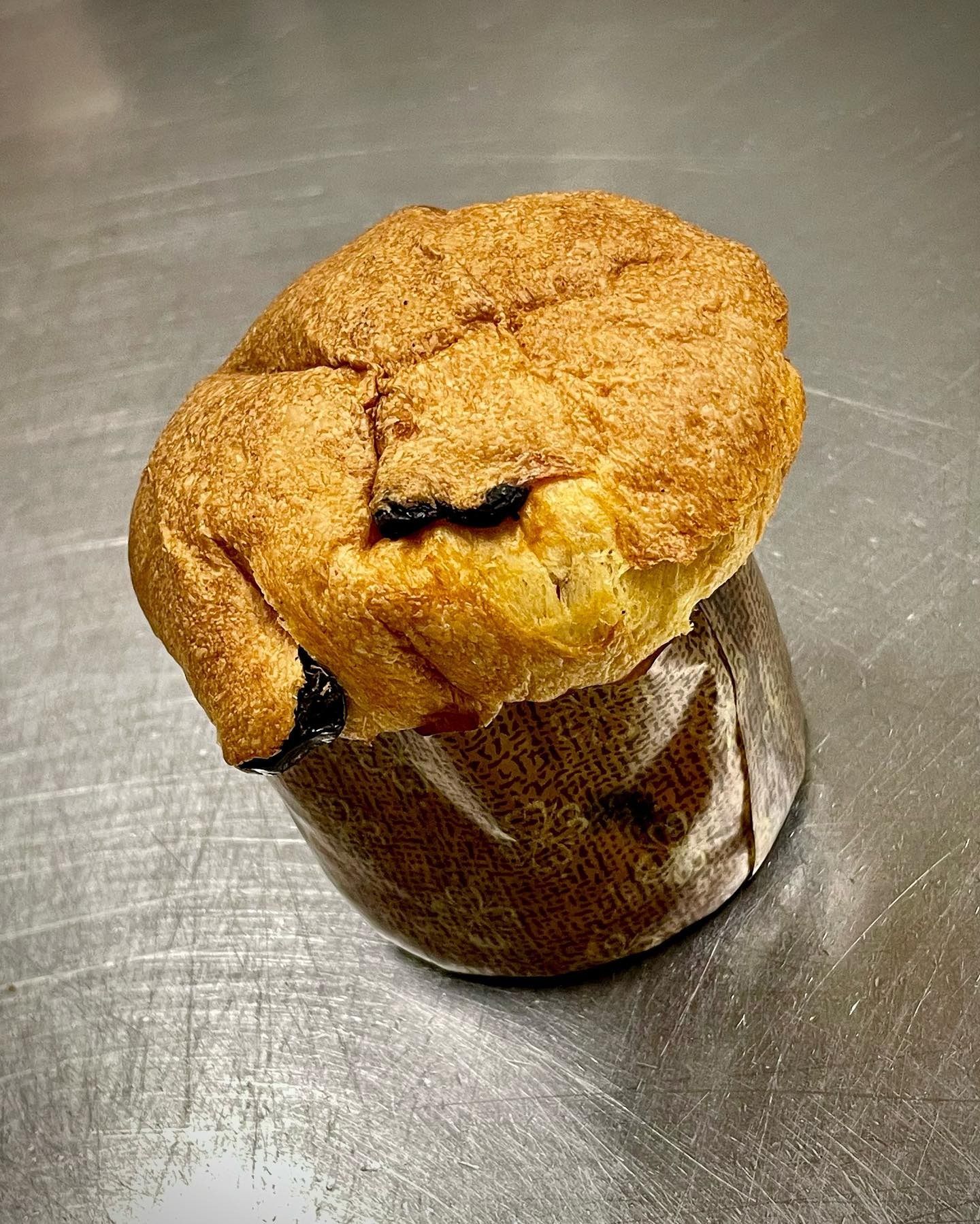 Un muffin dorato con la superficie impolverata, che esce da un bicchiere di carta marrone accartocciato, è appoggiato su una superficie metallica riflettente.