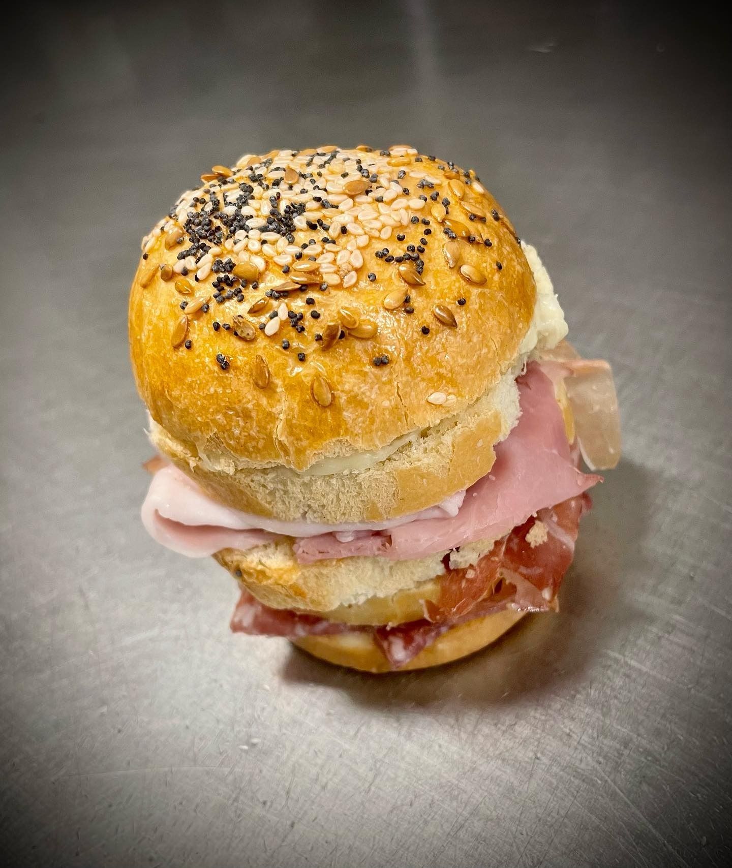 Un piccolo panino con prosciutto e salame, disposto su un panino con semi e appoggiato su una superficie in acciaio inox.