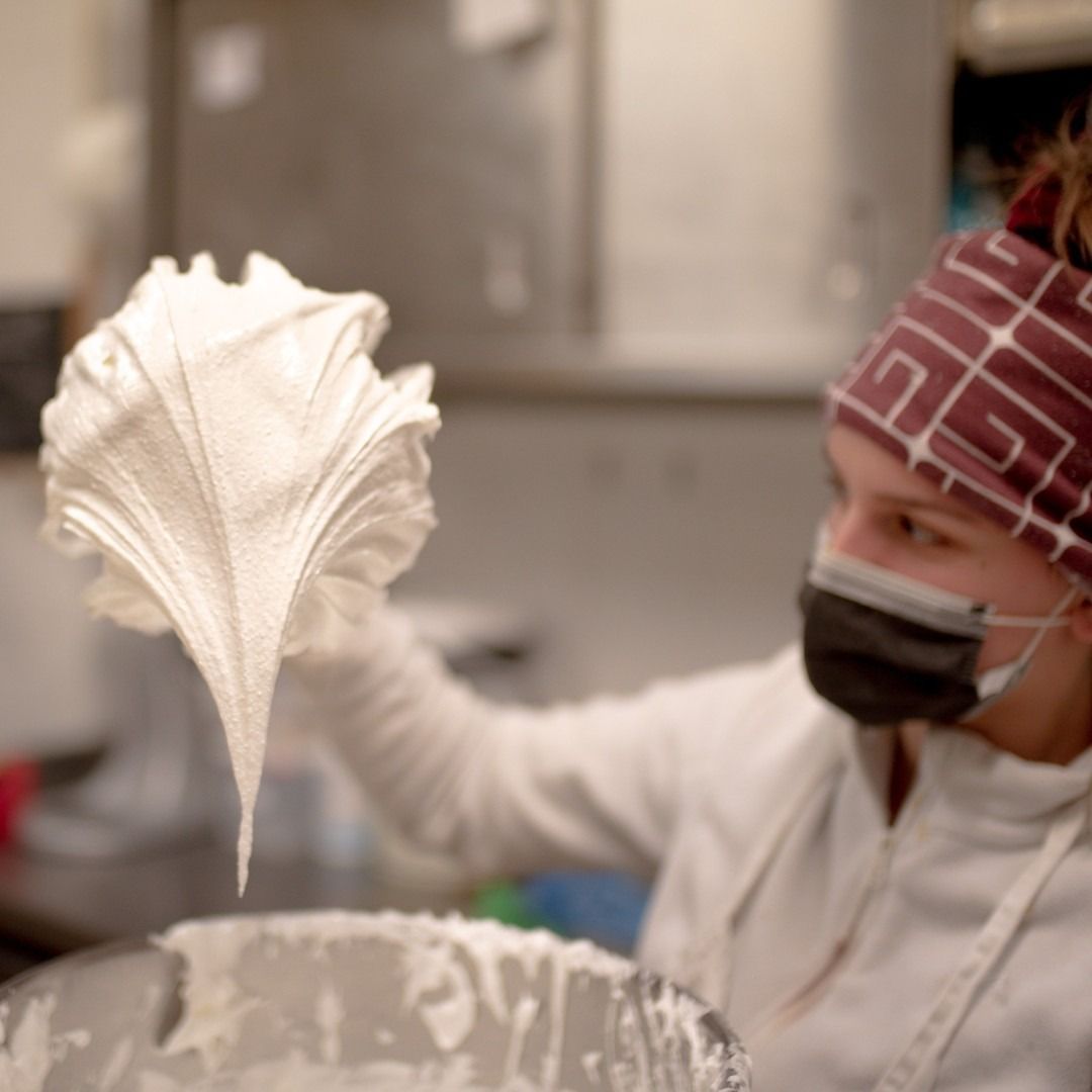 Una persona in cucina tiene in mano una cucchiaiata di meringa bianca, indossando una mascherina e un berretto.