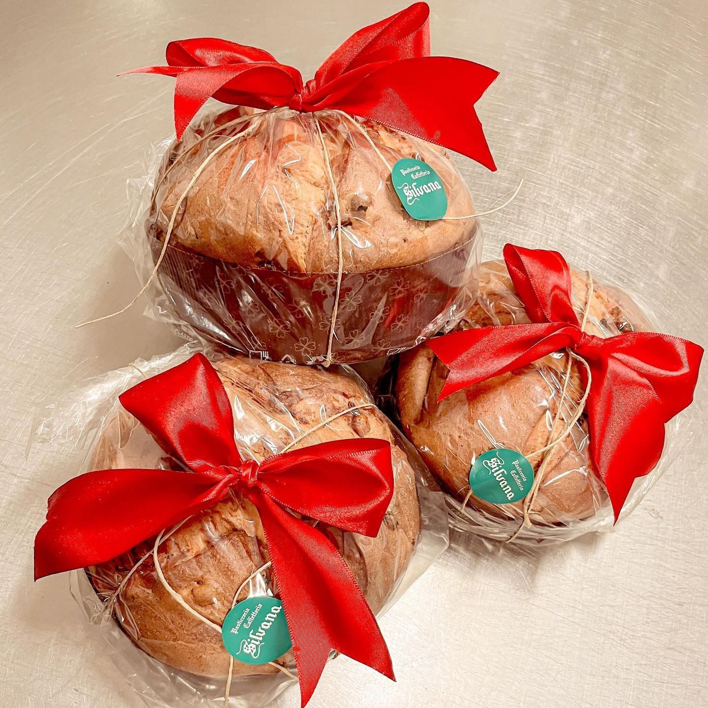 Tre prodotti da forno rotondi, avvolti in cellophane trasparente e legati con fiocchi rossi. Ognuno ha una piccola etichetta verde.