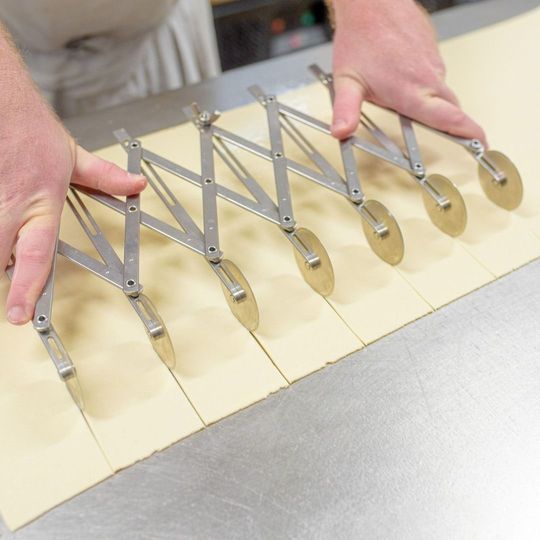 Mani che utilizzano un tagliapasta regolabile con più lame per dividere l'impasto in strisce uniformi su una superficie in acciaio inossidabile.