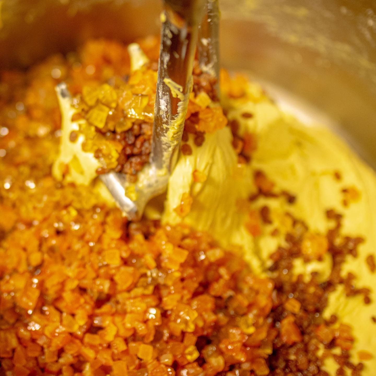 Impasto giallo mescolato con frutta candita arancione in una ciotola di metallo, con la frusta a foglia visibile.