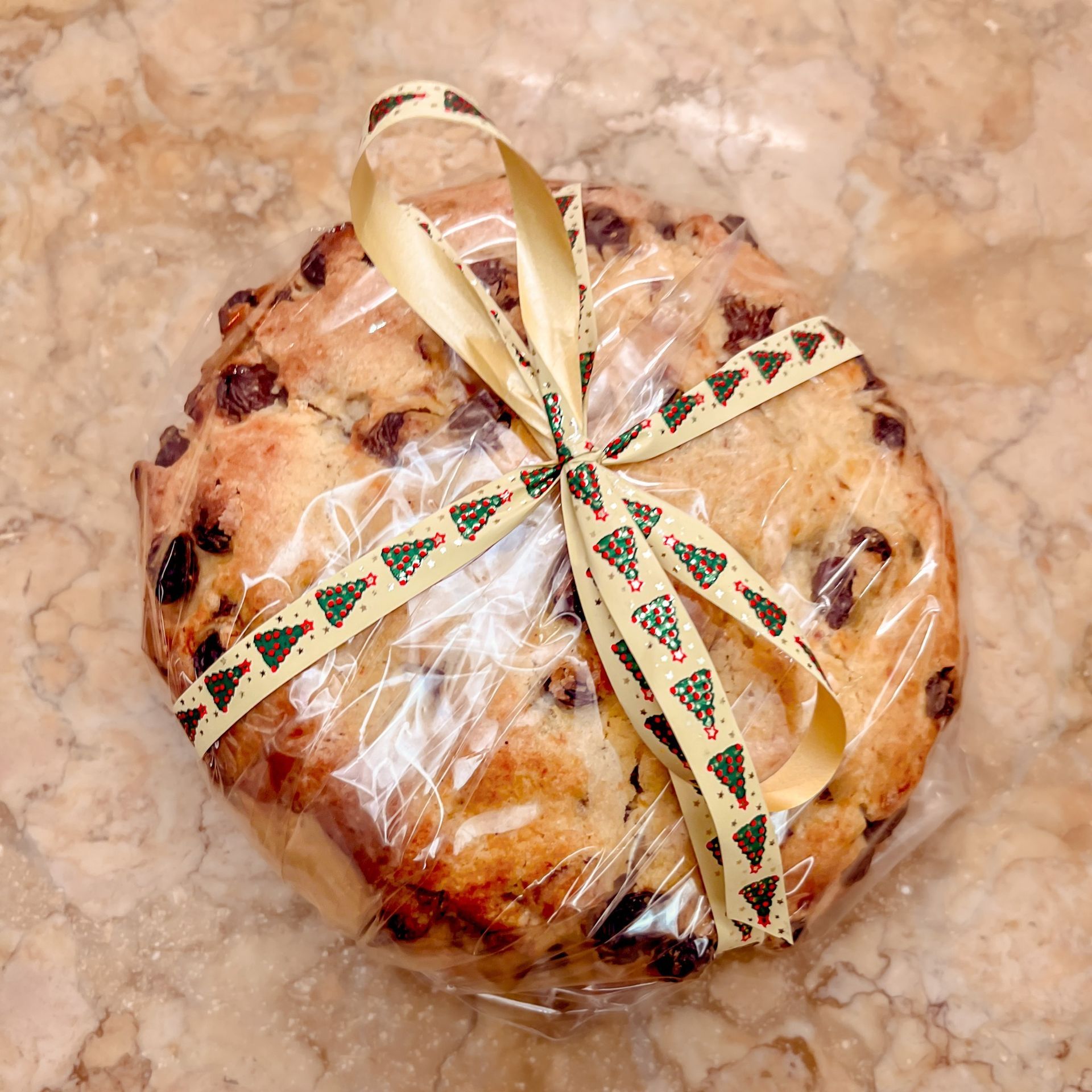Un dolce rotondo, ricoperto di gocce di cioccolato, avvolto in cellophane e legato con un nastro decorato con alberi di Natale, appoggiato su una superficie di marmo.