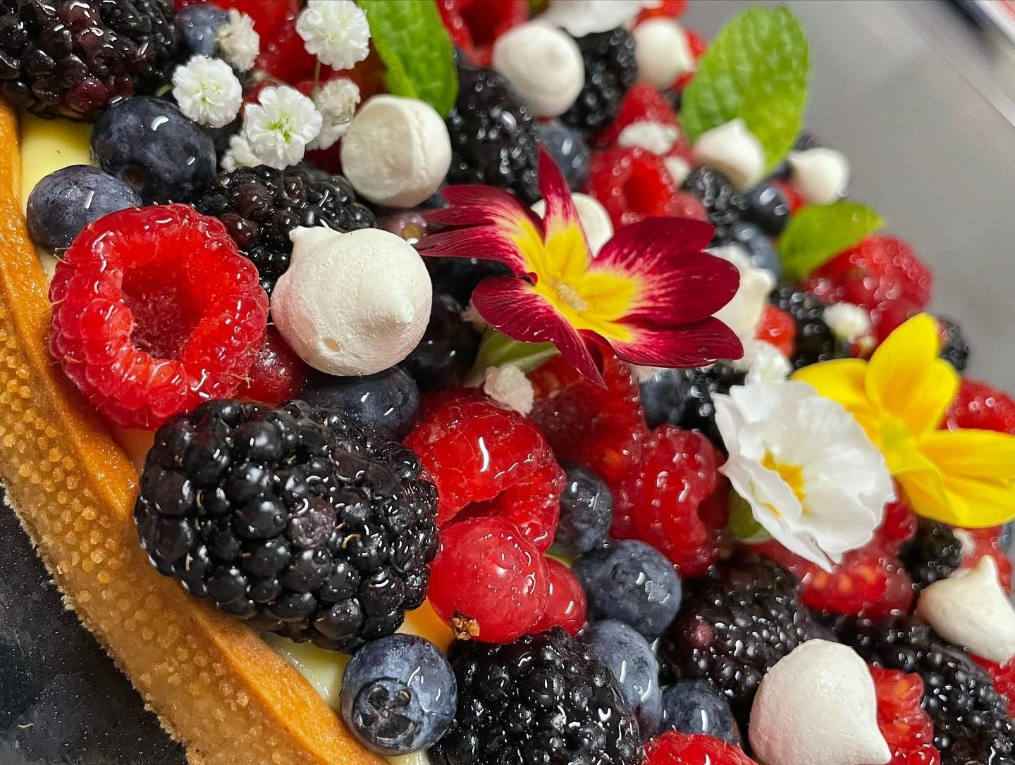 Primo piano di una crostata ai frutti di bosco guarnita con lamponi freschi, more, mirtilli, meringhe e fiori commestibili.