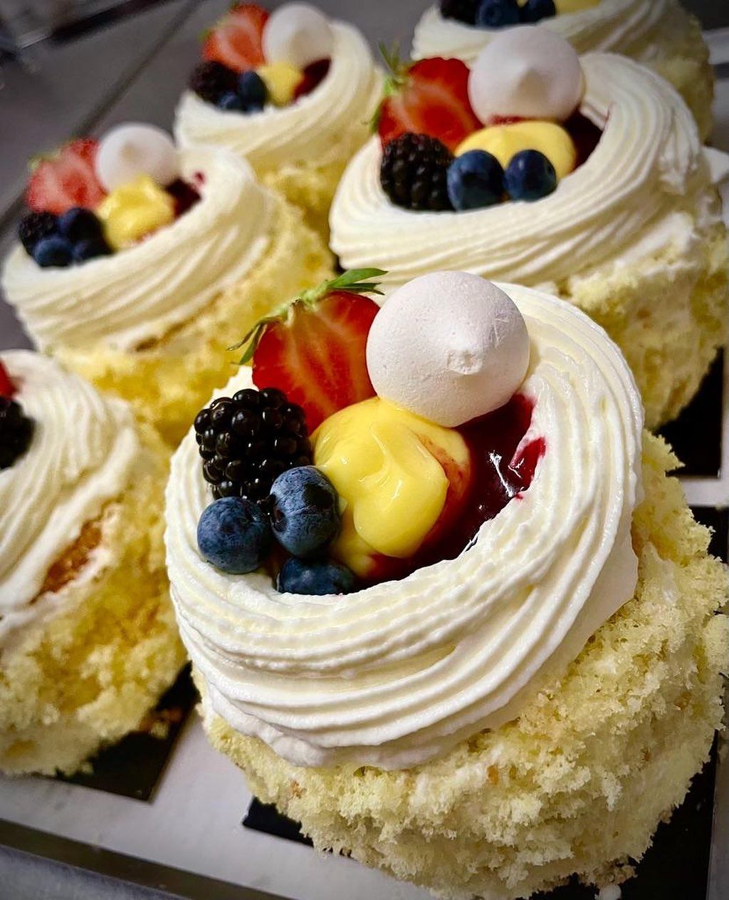 Primo piano di diverse mini torte spugnose ricoperte di panna montata, frutti di bosco freschi e un ciuffo di crema pasticcera gialla.