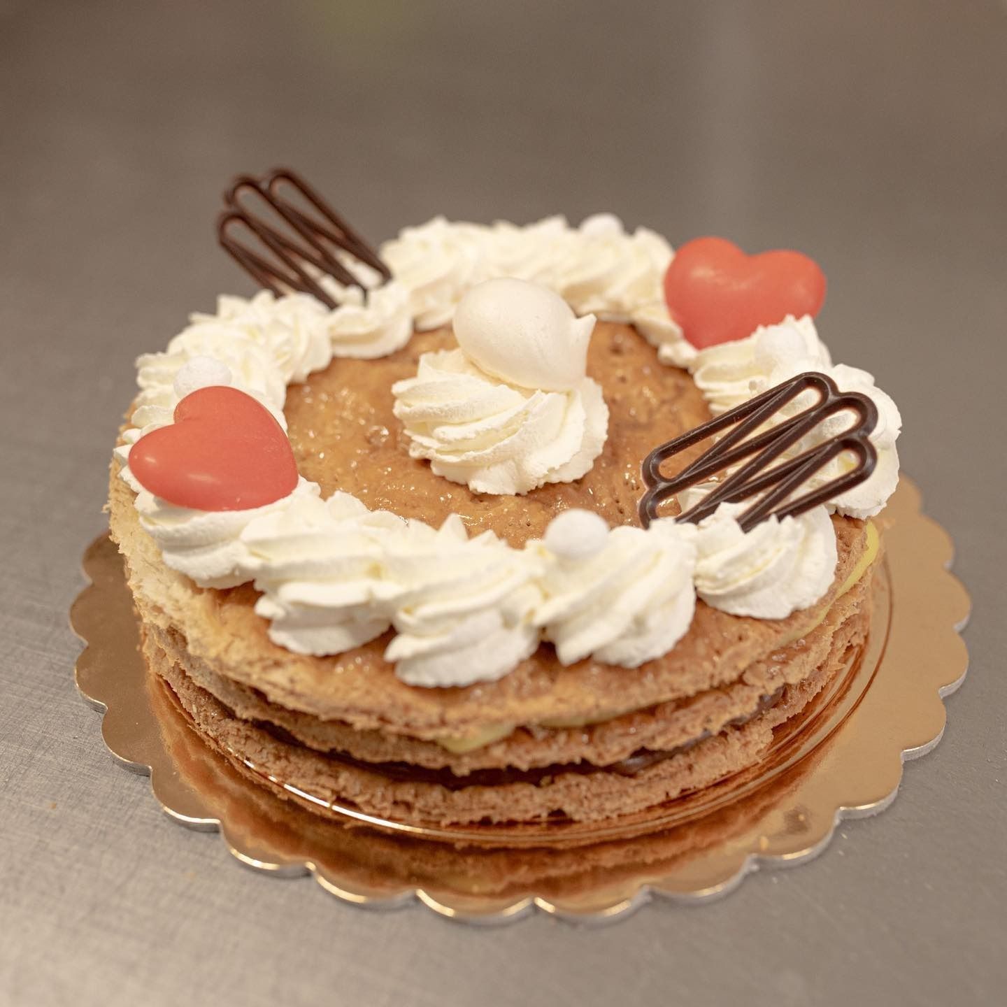 Una torta dolce a strati con panna montata, decorazioni di cioccolato e caramelle a forma di cuore rosso su un centrino di carta dorato e smerlato.
