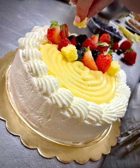 Torta con crema pasticcera gialla al centro, guarnita con frutti di bosco e glassa a spirale, su un piatto dorato smerlato. Qualcuno cosparge la superficie con briciole di torta.