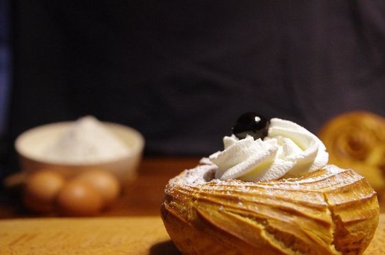 Primo piano di un dolce con panna montata e una ciliegia, con farina, uova e un altro dolce sfocati sullo sfondo.