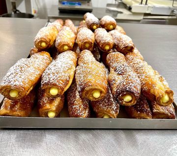 Un vassoio di pasticcini dorati e cilindrici, ripieni di crema gialla e spolverati con zucchero a velo, probabilmente utilizzato in una pasticceria.