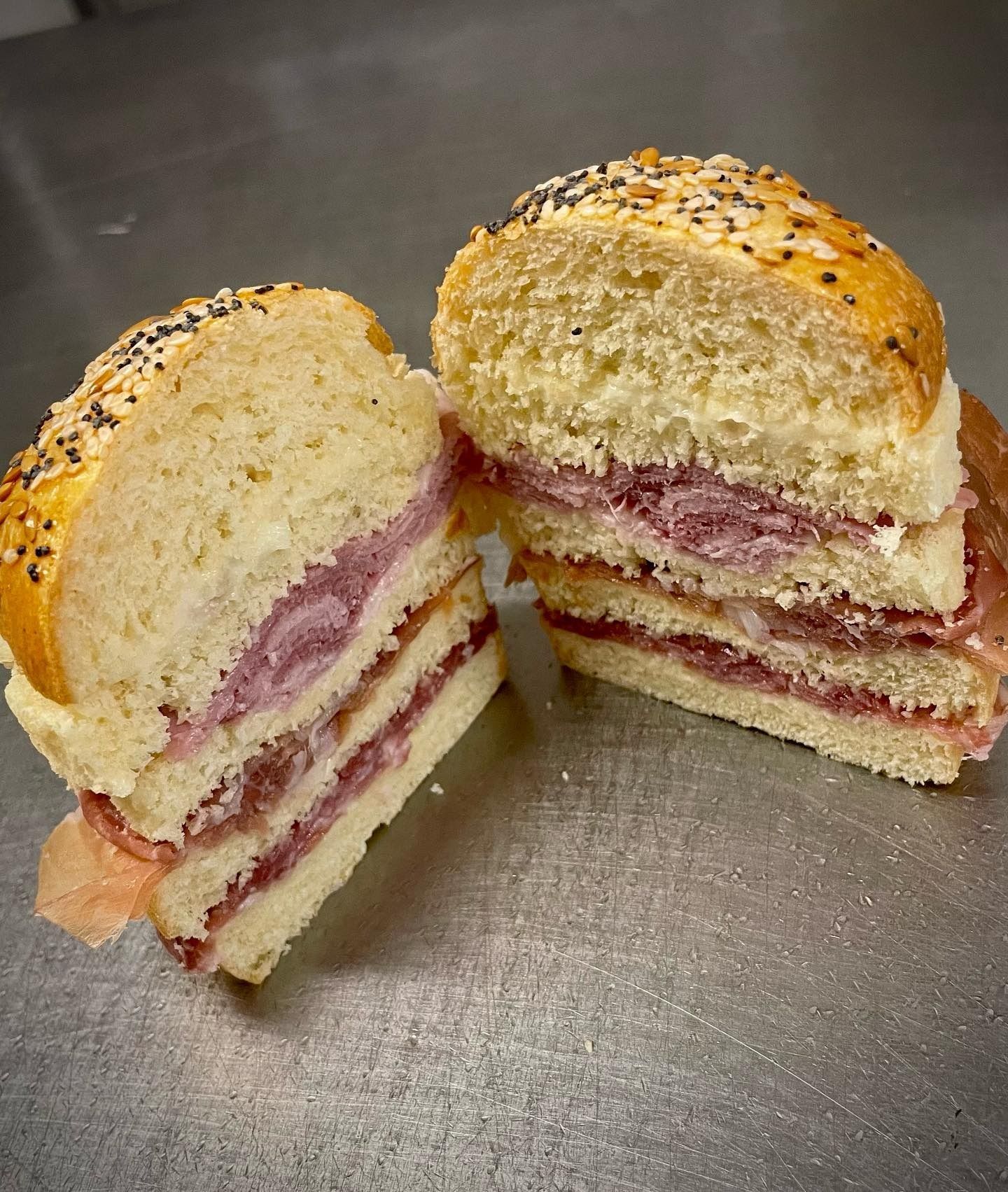 Un panino affettato su un panino con semi, ripieno di strati di carne e una salsa chiara, appoggiato su una superficie metallica.