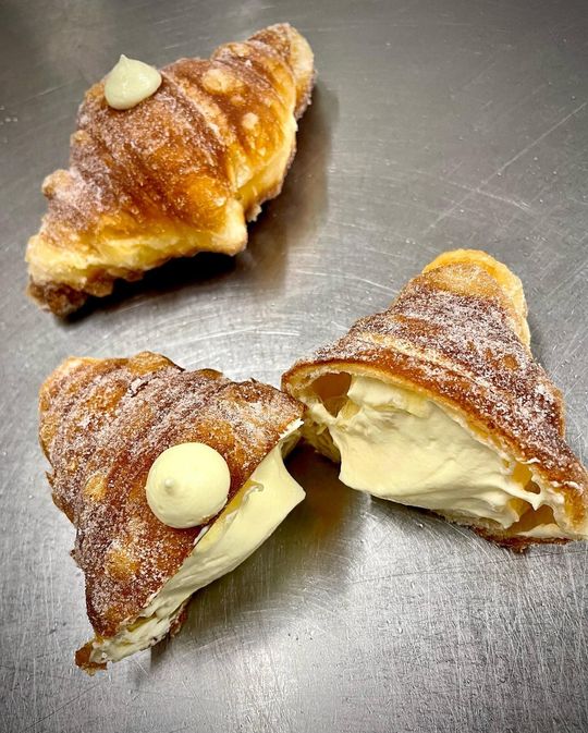 A puff pastry croissant, opened to reveal a creamy filling. The outside is dusted with sugar and garnished with a tuft of cream.