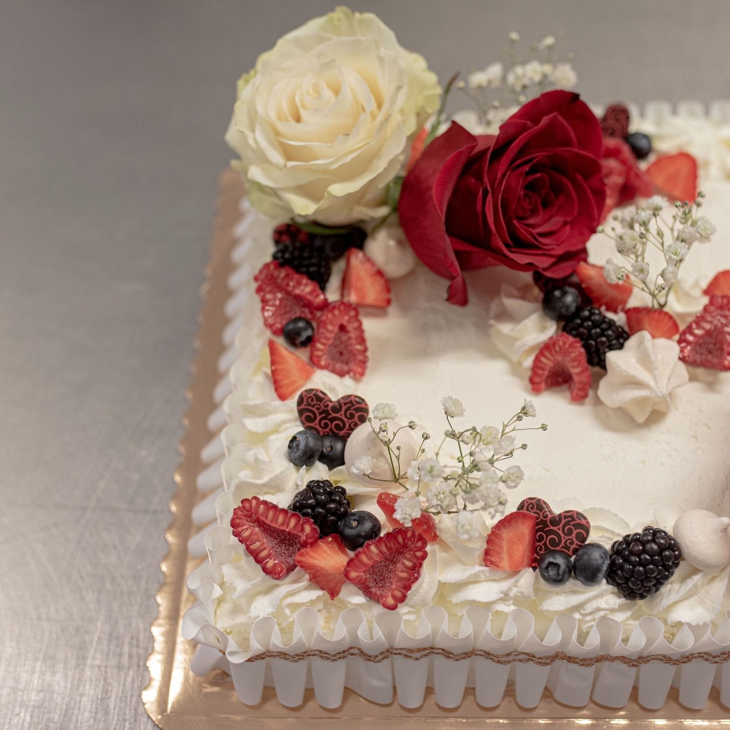Torta quadrata decorata con glassa bianca, frutti di bosco freschi e rose. La torta è adagiata su un vassoio dorato.
