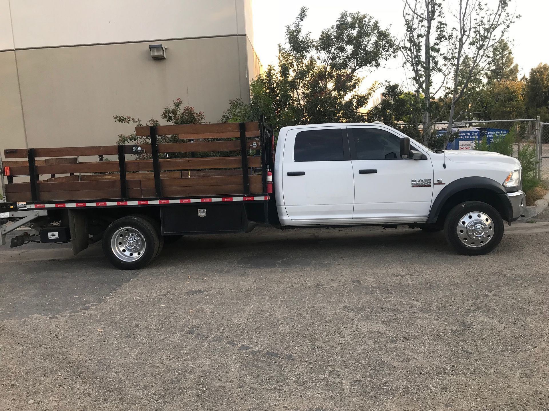 White Ram 5500 flatbed truck parked on asphalt. Brown wooden bed, black trim, chrome wheels, and trees in background.