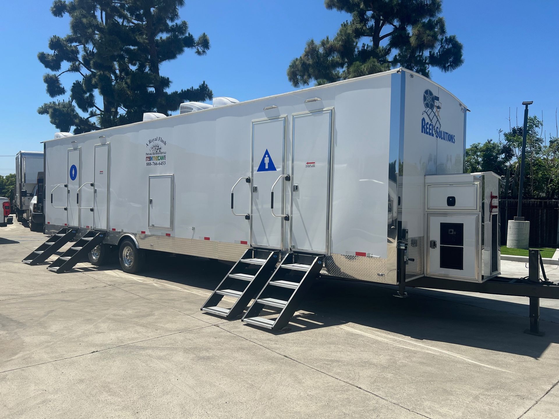 White trailer with multiple access doors and ramps, parked outdoors on concrete.
