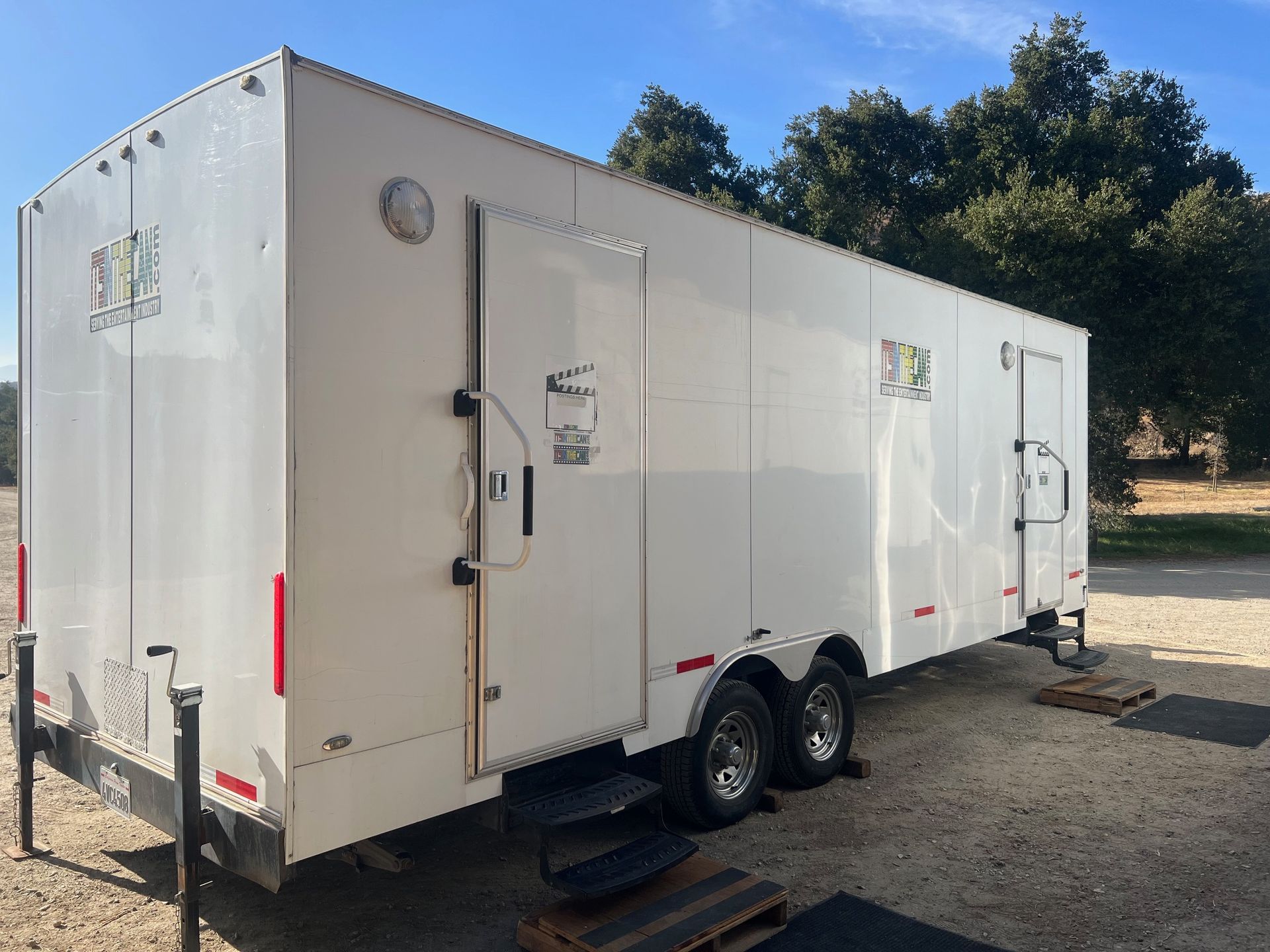 White portable restroom trailer with two entry doors, sitting on a gravel lot under a blue sky.