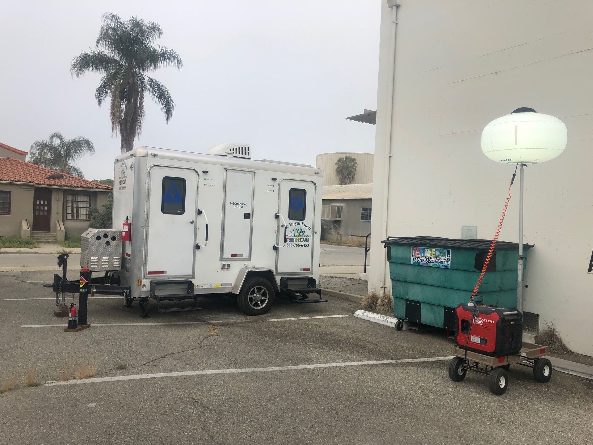 White portable restroom trailer and generator in a parking lot next to a building and dumpster.