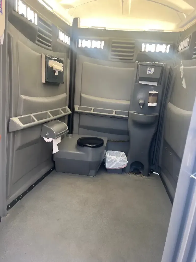 Interior of a gray portable toilet. Features include a toilet seat, hand sanitizer dispenser, and vents.