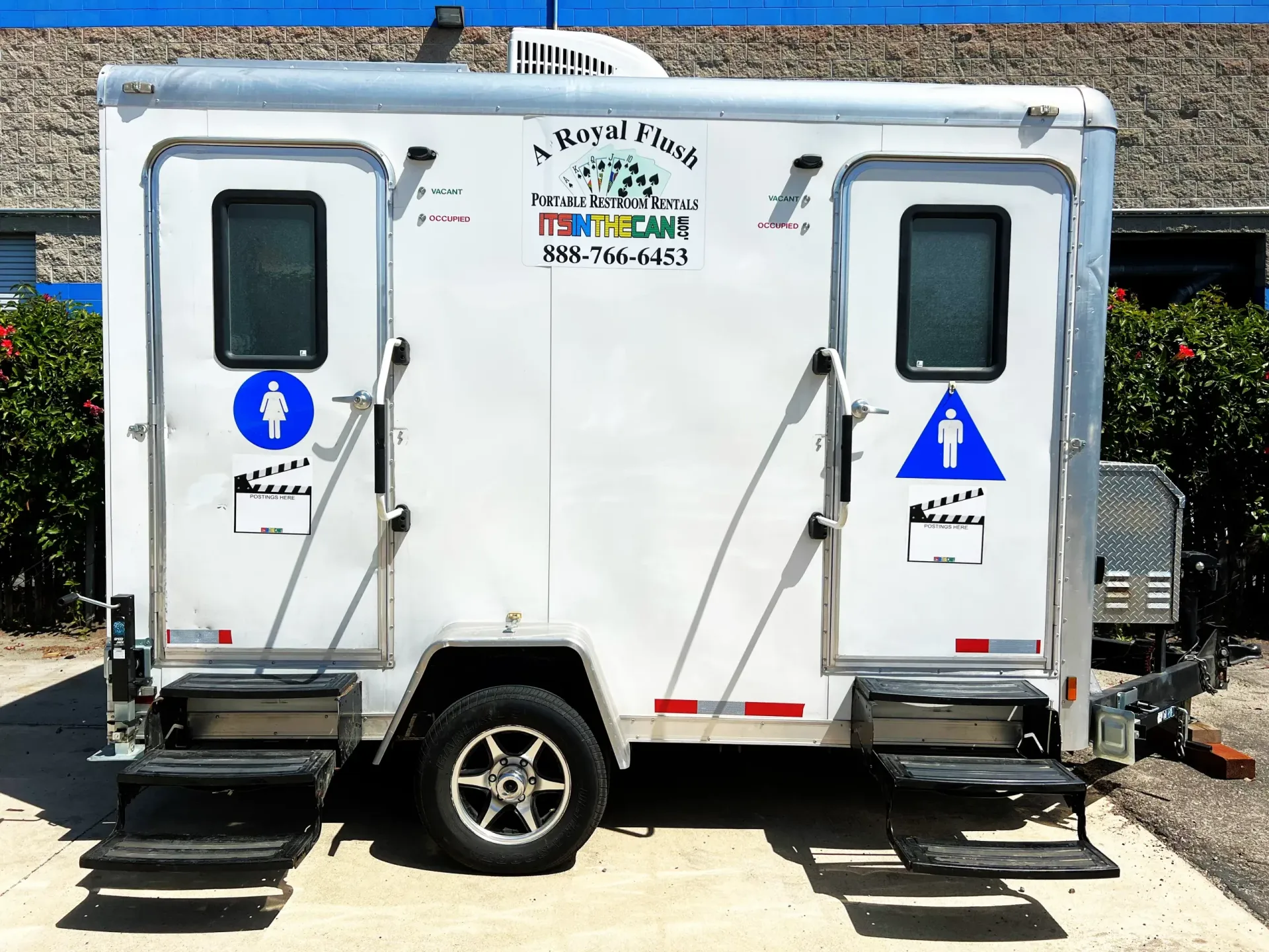 Portable restroom trailer with two entrances. White with blue signage and steps.