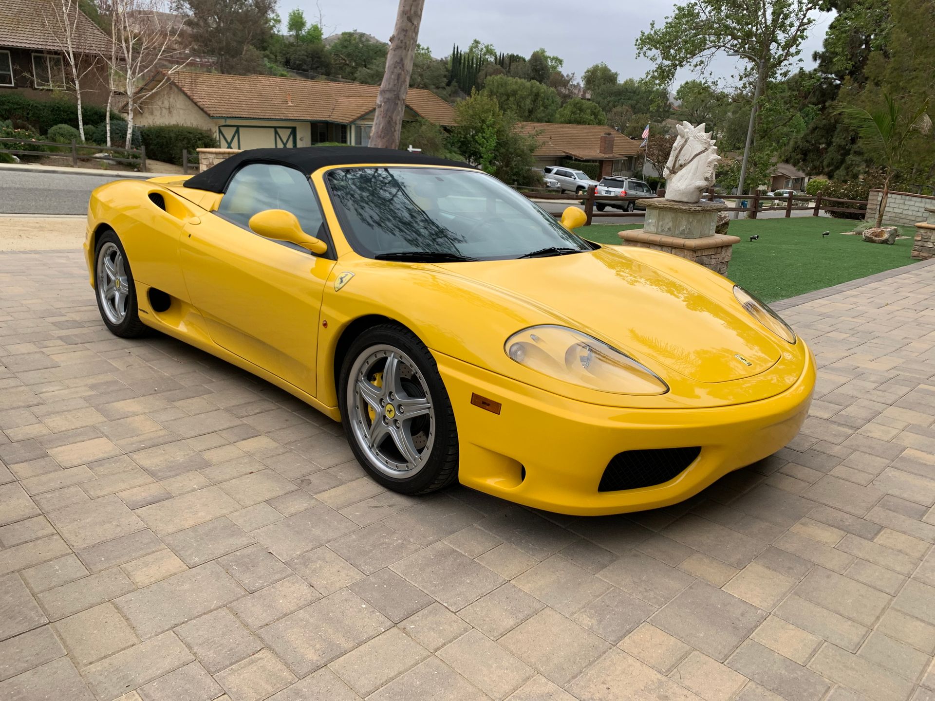 Yellow Ferrari convertible parked on a brick driveway, black top down, houses in the background.