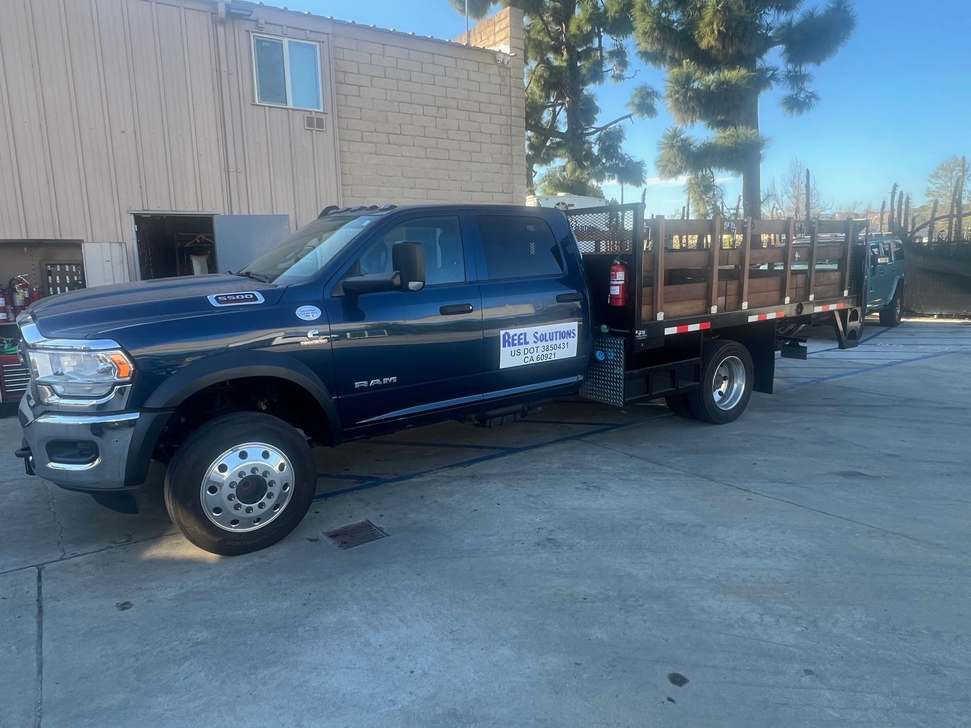 Blue flatbed truck parked in front of a building with trees; the truck has a company logo on the door.