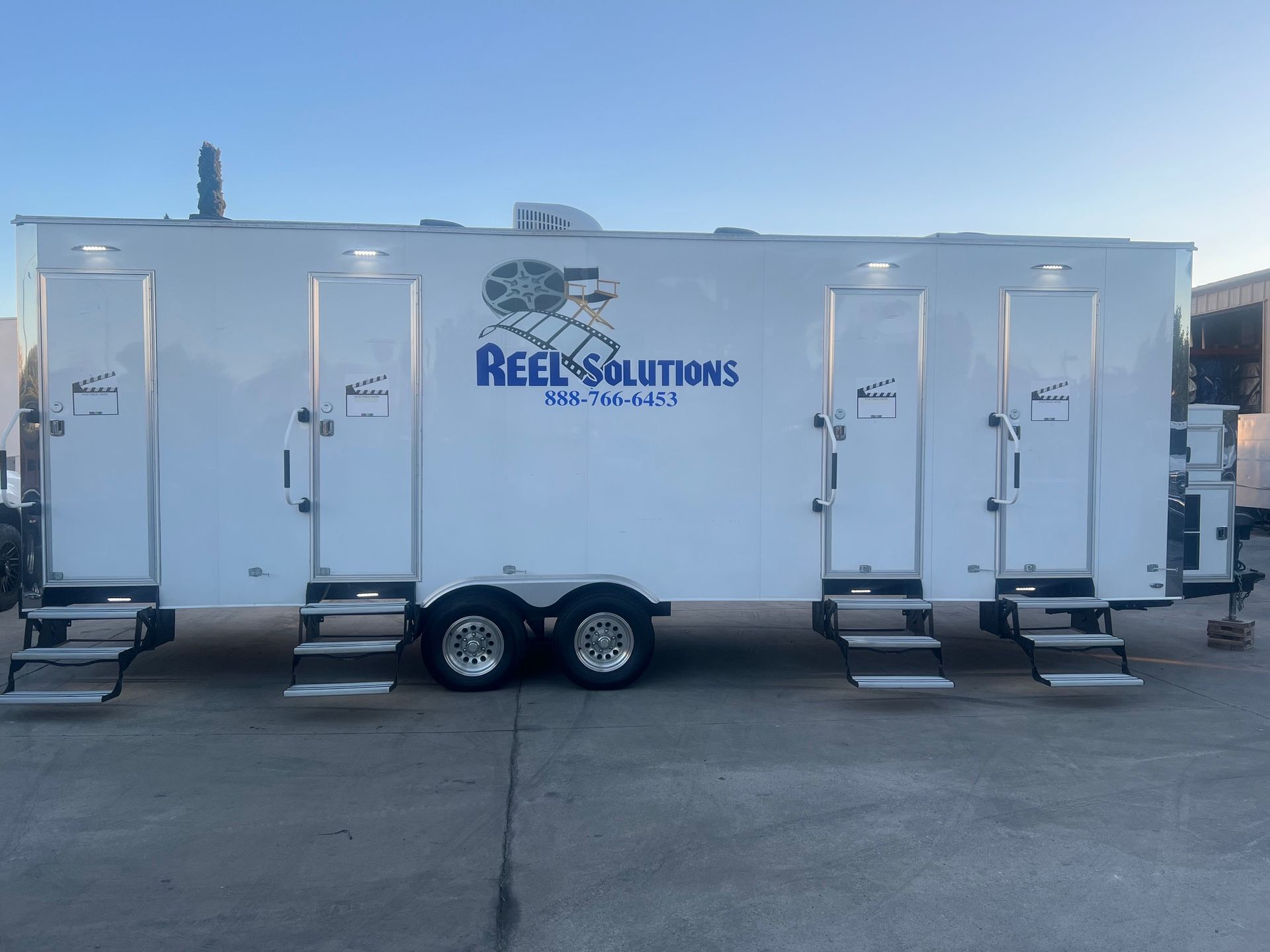 White mobile restroom trailer with steps, REEL SOLUTIONS logo on side.