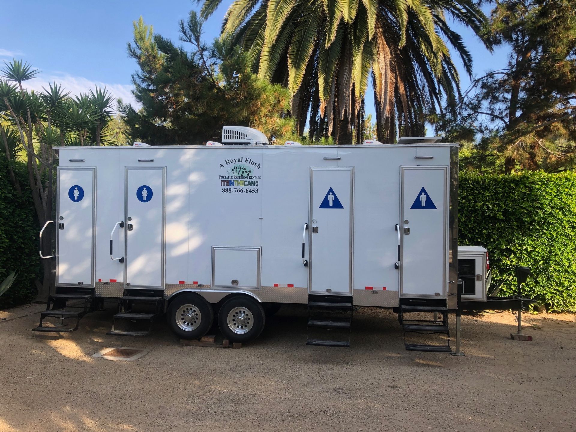 Portable restroom trailer with multiple stalls, parked outdoors near foliage.