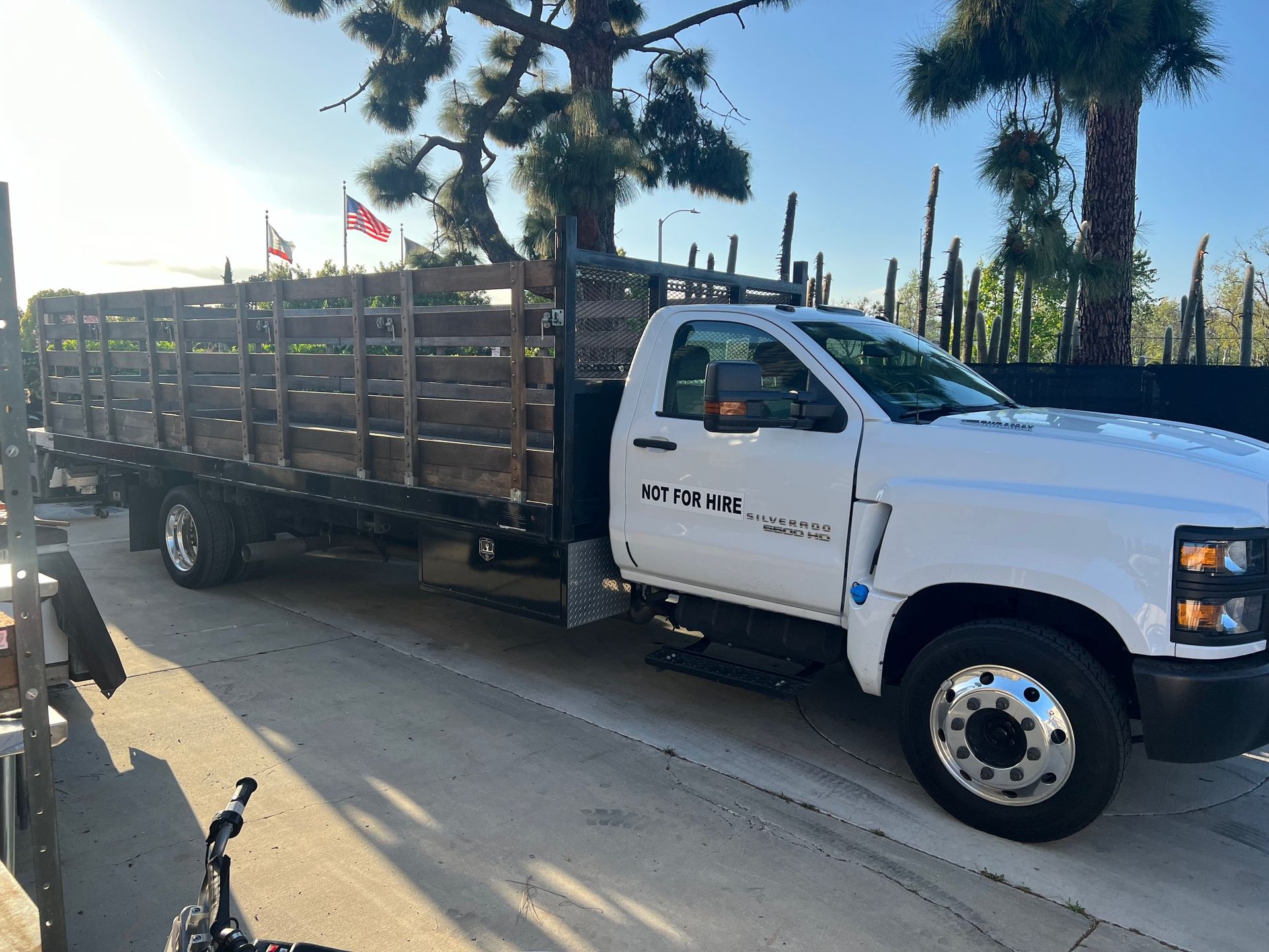 White flatbed truck parked on a paved driveway in front of a building with trees.