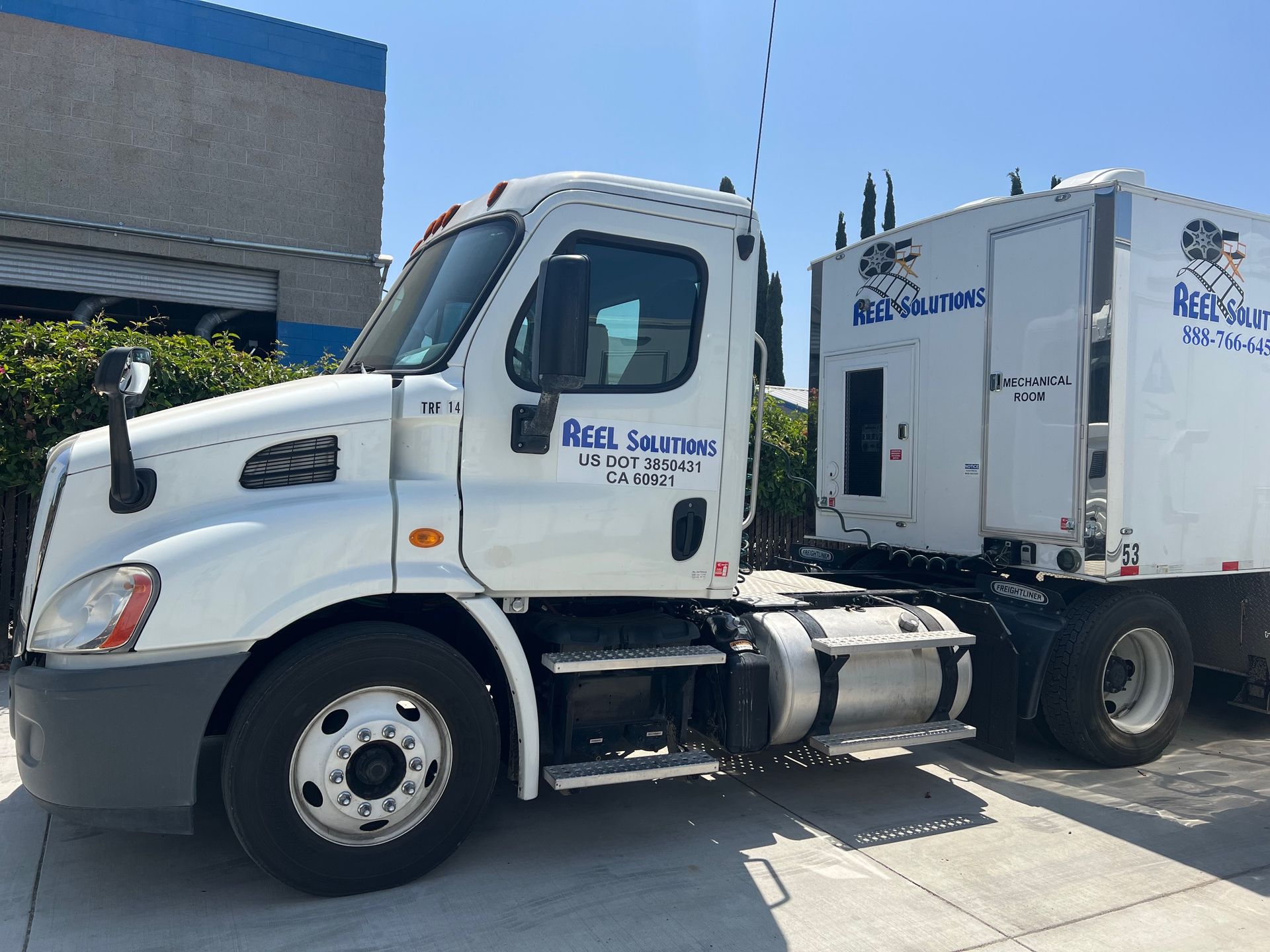 White semi-truck with trailer, blue lettering, parked outside a building on a sunny day.