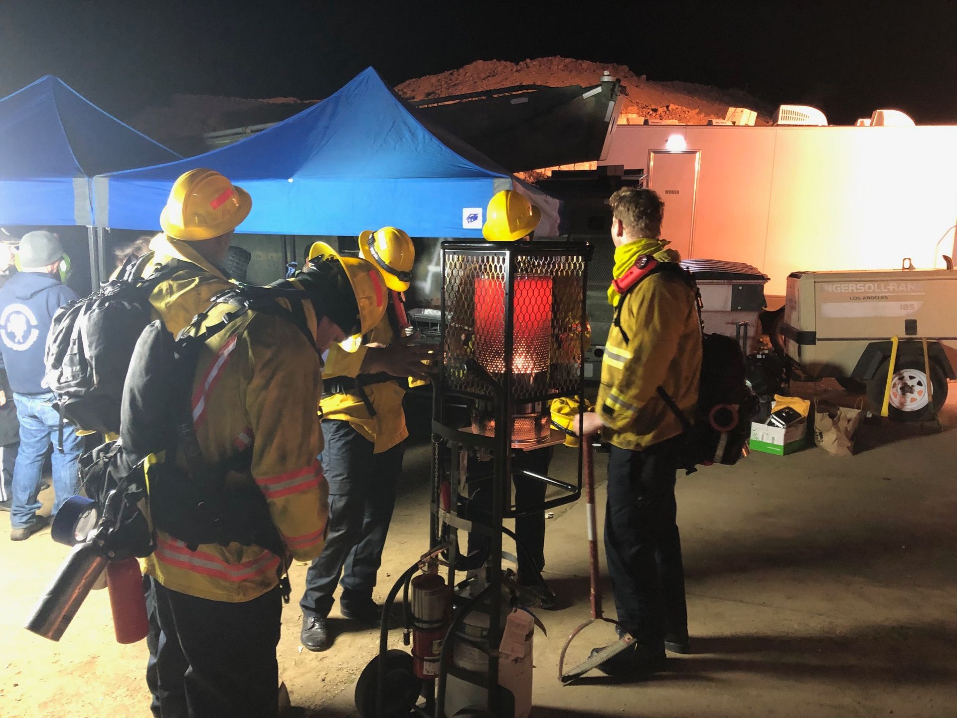 Firefighters in yellow gear examining equipment at night. Blue tent in background.