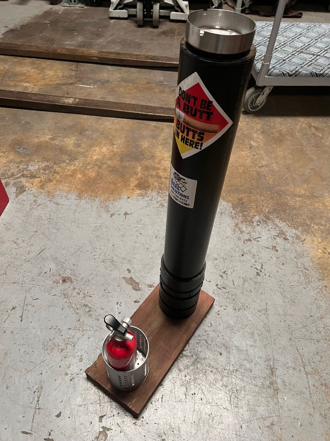 A tall black cylindrical object with a silver top sits on a wooden base next to a small red fire extinguisher.