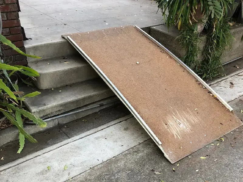 Ramp made of brown material with metal edges, set against gray concrete steps leading to a building entrance.