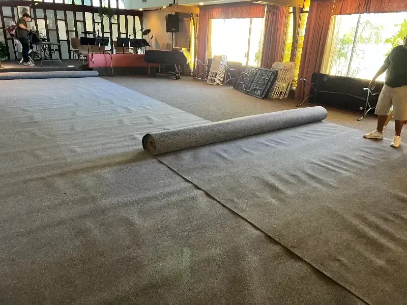 Person rolling out new gray carpet in a large room, likely for an event space.