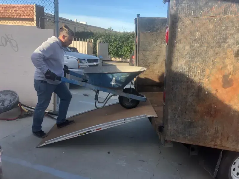 Man using a wheelbarrow to unload into a dumpster, on a ramp. Outdoors.