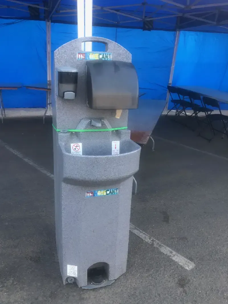 Gray portable handwashing station with soap, paper towels, and foot pump, outdoors under a blue canopy.