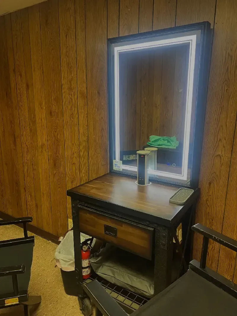 Vanity with a lit mirror. Brown wood paneling, black and brown furniture with a drawer and shelf.
