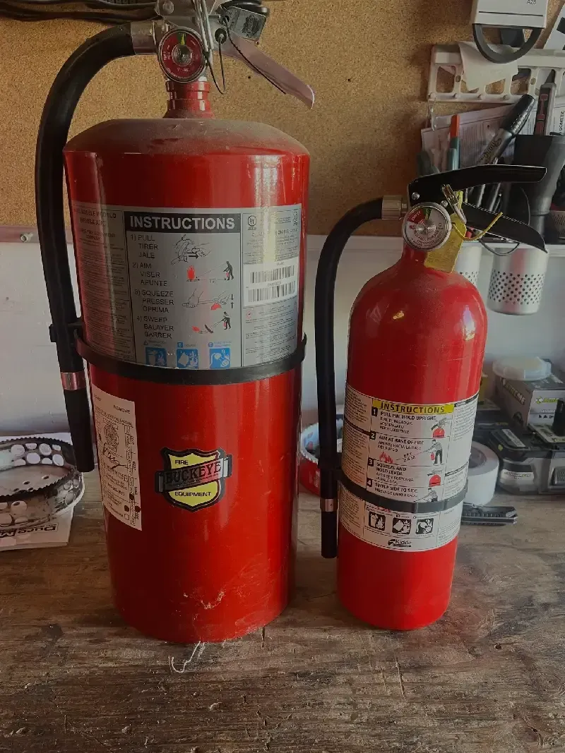 Two red fire extinguishers of different sizes, sitting on a cluttered surface.