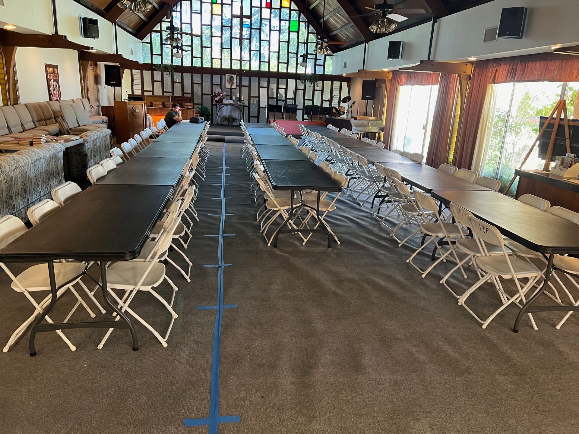 Interior with long tables and white chairs set up for an event. Stained glass window in background.