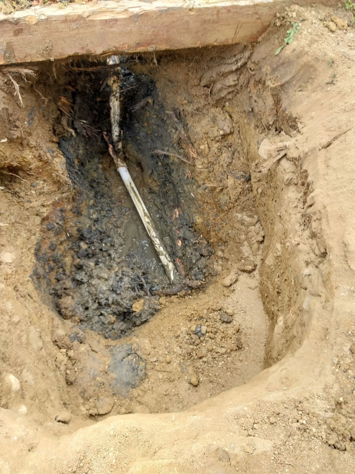 A hole dug in dirt reveals a damaged pipe, surrounded by dark, possibly contaminated soil.