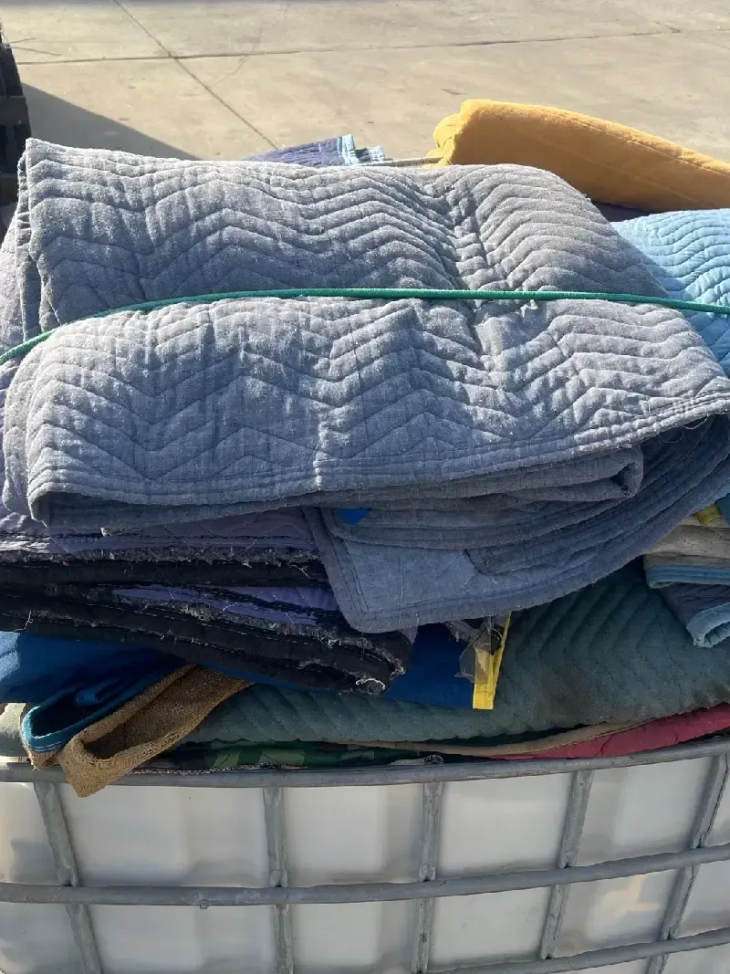 A stack of folded, quilted moving blankets, bound with a green strap, on a white bin.