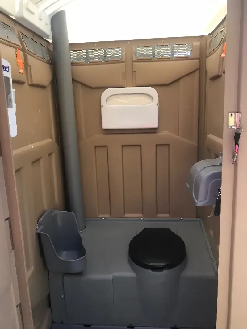 Interior of a portable toilet. Beige walls and floor with a black toilet seat, urinal, and paper towel dispenser.