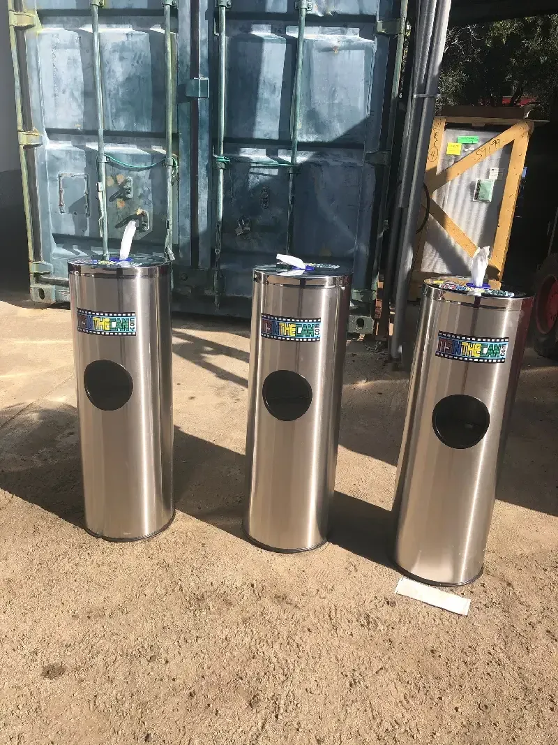 Three tall, silver, cylindrical trash cans on a dirt surface. Each has a black circular opening and a logo.