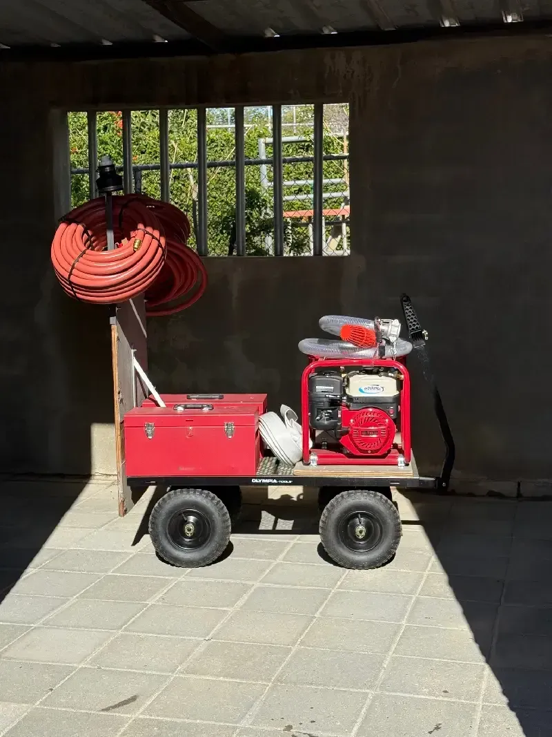 Red utility cart with hose reels, gas engine, and box. Parked near a window.