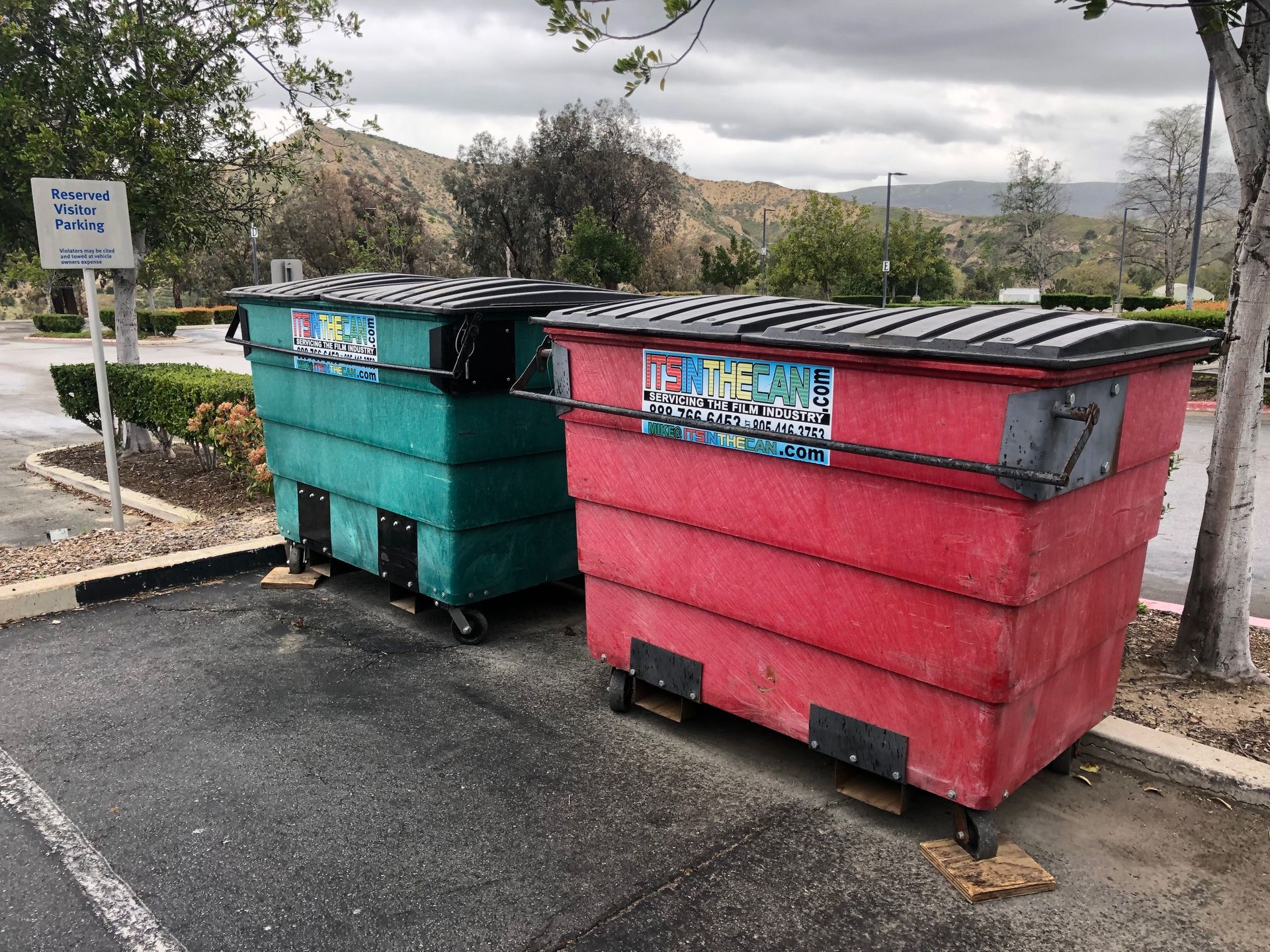 Two large dumpsters, one teal, one red, on a gray asphalt surface, possibly in a parking area with hills in the background.